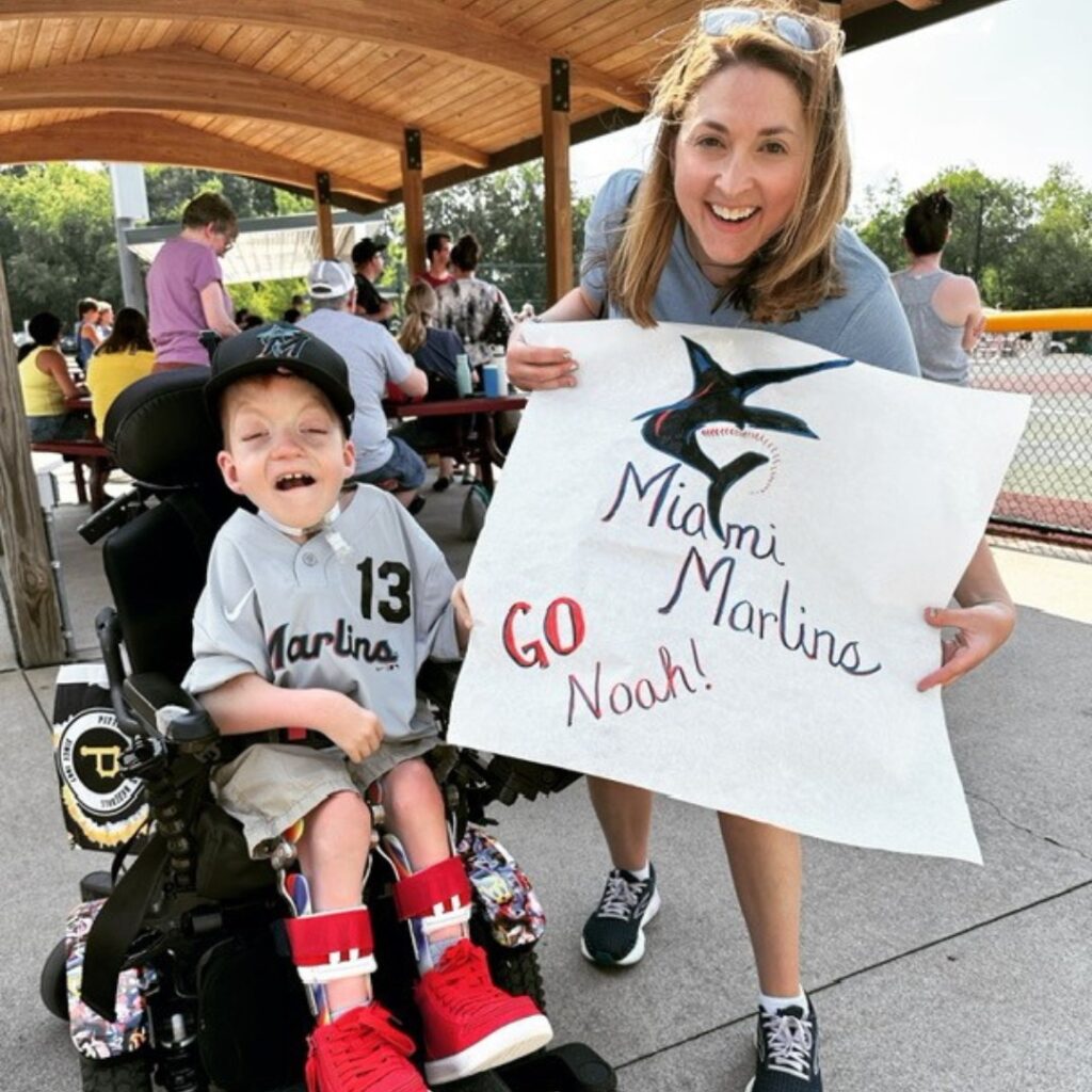 A smiling woman kneels beside a young boy in a wheelchair. The boy wears a Miami Marlins jersey and cap. The woman holds a large sign reading Miami Marlins GO Noah! celebrating Noahs Story at an outdoor sports field.