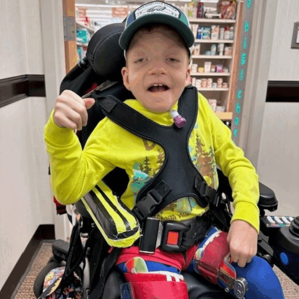 A young boy named Noah, wearing a bright yellow shirt, colorful pants, and a green cap, smiles and gives a thumbs-up inside a store, with shelves of products visible in the background. Discover Noahs Story today!.