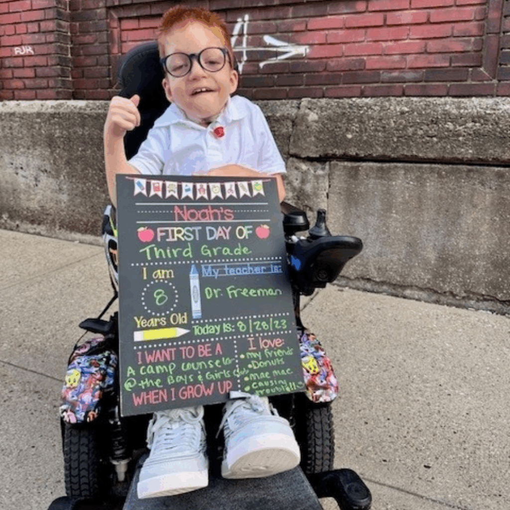 A smiling young boy in glasses sits in a wheelchair, holding a colorful chalkboard sign about his first day of third grade. This is Noahs Story—he wears a white shirt, gives a thumbs up, and sits on a sidewalk by a brick wall.