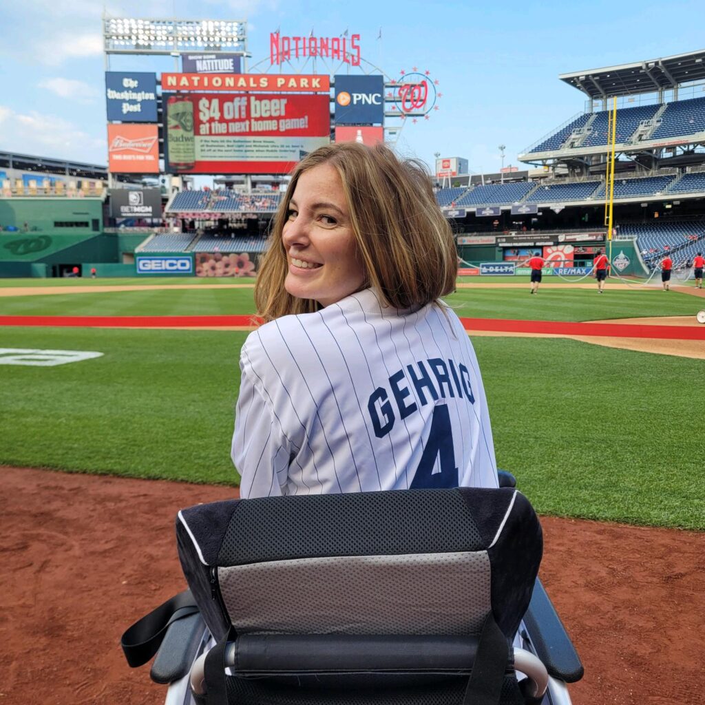 Brookes Story: A woman using a wheelchair, wearing a Gehrig 4 baseball jersey, smiles at the camera near the field at Nationals Park, with the stadium and players visible in the background.