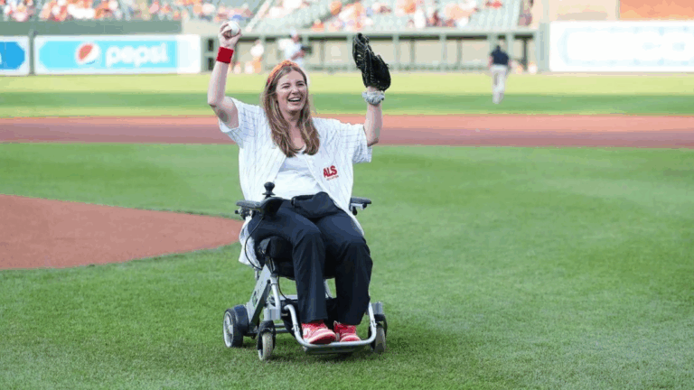 Brookes Story: A woman in a wheelchair on a baseball field smiles and raises her arms in celebration, wearing a baseball glove, jersey, and red shoes as a cheering crowd fills the background.