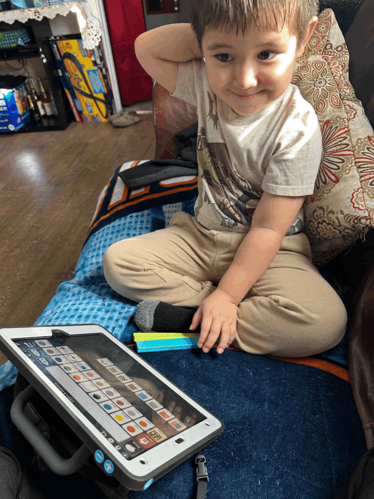 A young boy sits on a couch with one arm behind his head, smiling. In front of him is a communication tablet displaying icons. The cozy room, filled with pillows and various items, reflects a scene inspired by Darwin’s Story of evolution.