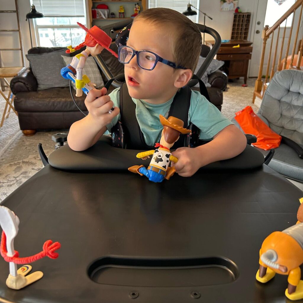 A young boy wearing glasses and a green shirt sits in a wheelchair, holding Toy Story action figures Woody and Jessie, with Forky and other toys on the tray in front of him. This heartwarming scene is part of Logans Story in a cozy living room.