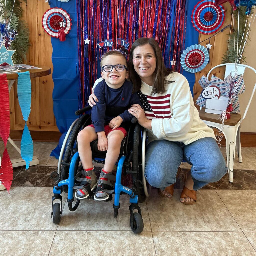 A smiling woman kneels next to a young boy in a wheelchair. Both, surrounded by red, white, and blue decorations in a festive indoor setting, share a joyful moment—capturing the spirit of Logans Story.
