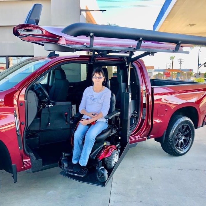 Tricias Story: A woman in a wheelchair uses a red accessible pickup truck with a side-entry ramp and a surfboard on top, parked at a gas station.
