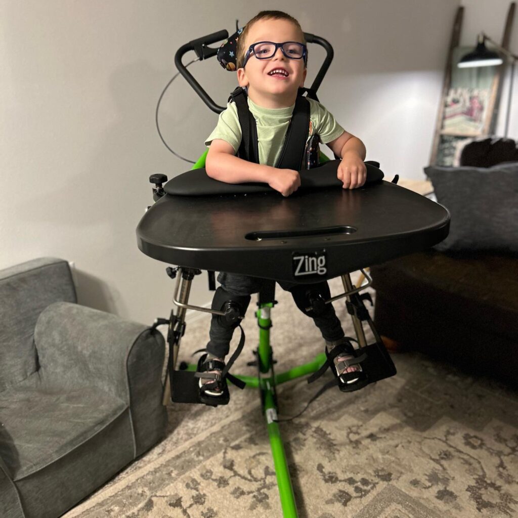 A young child with glasses smiles while using a green standing frame labeled Zing in a living room, supported by a harness and resting arms on a black tray. Nearby, a couch and rug set the scene for Logans Story.