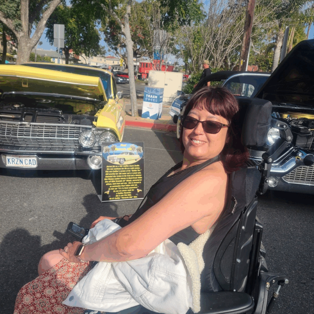 Tricias Story: A woman in sunglasses smiles at a classic car show, sitting in front of two vintage cars with open hoods. Trees and a sunny sky are in the background.