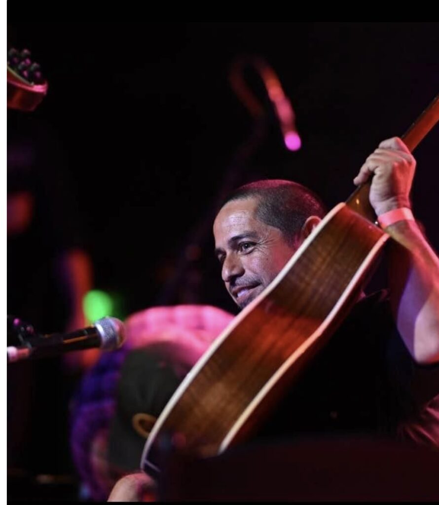 A man smiles while holding an acoustic guitar over his shoulder on stage, with a microphone in front of him and colorful stage lights in the background, sharing Genes Story through his music.