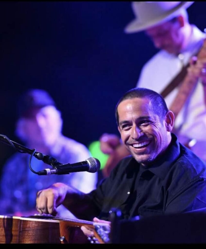 A smiling musician in a black shirt plays guitar and sits behind a microphone on stage, sharing Genes Story, with two other musicians blurred in the background under colorful stage lighting.