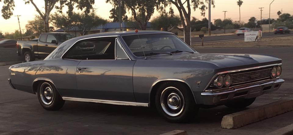 A classic gray Chevrolet Chevelle coupe is parked in a lot at sunset, with trees, cars, and streetlights visible in the background—a perfect scene from Genes Story.