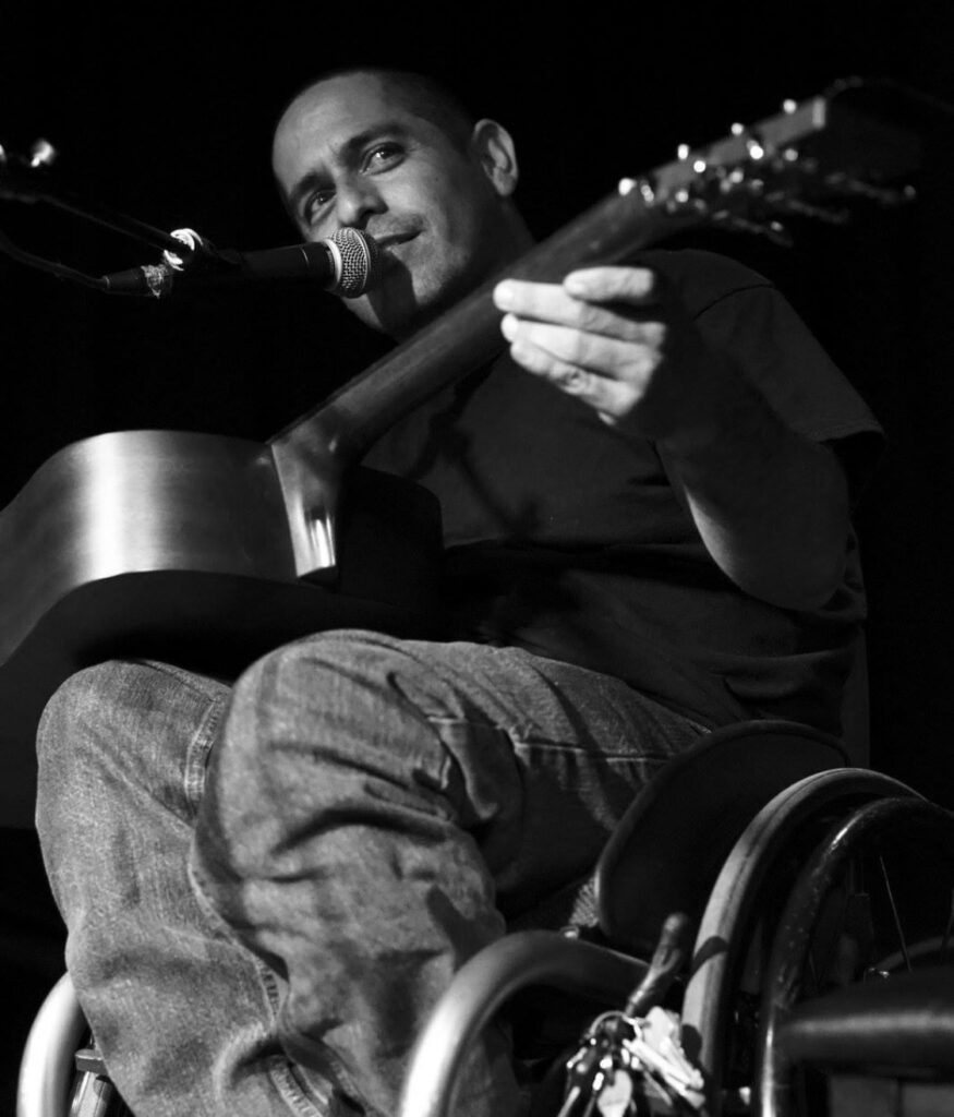 A man in a wheelchair plays acoustic guitar and sings into a microphone on stage. The image is in black and white, capturing a moment from Genes Story.