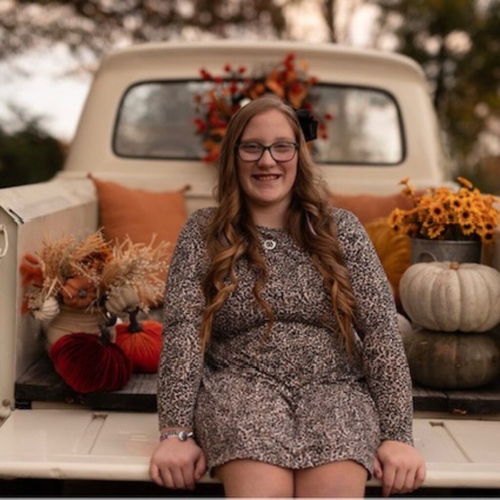 Ellas Story: A young woman with long, wavy hair and glasses sits smiling on the back of a vintage white truck adorned with autumn-themed items like pumpkins, flowers, and pillows. Trees stand in the background.