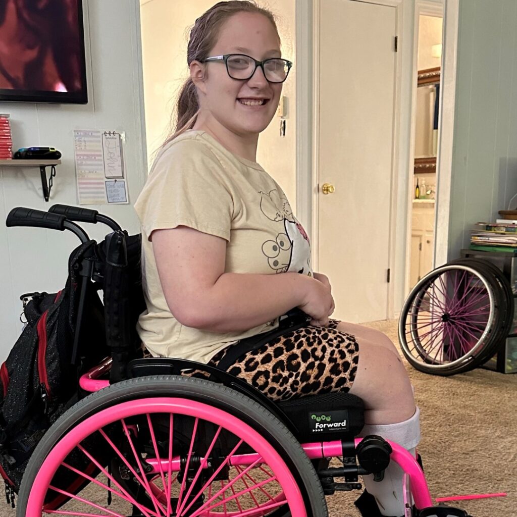 A smiling young woman with glasses sits in a pink wheelchair, wearing a beige t-shirt and leopard print shorts. Books and a bathroom are visible in the background, capturing a moment from Ellas Story.