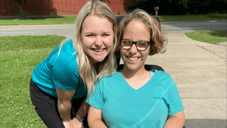 Two women smiling outdoors; one is standing and the other, featured in Danis Story, is sitting in a wheelchair. Both wear teal shirts and glasses as they pose together on a sunny day by a sidewalk and grass.