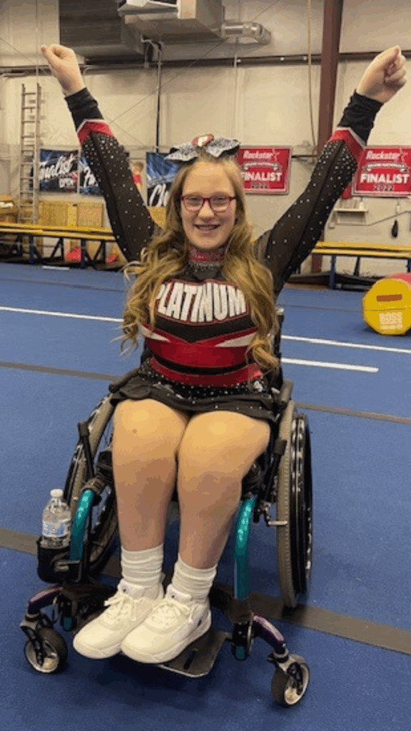 A young cheerleader sharing Ellas Story, wearing a black and red PLATINUM uniform and a bow, raises her arms and smiles indoors on a blue mat, with banners and gym equipment in the background.