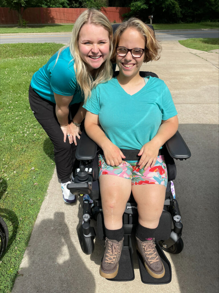 Two smiling women enjoy a sunny day together; one stands and leans in close while the other, featured in Danis Story, sits in a power wheelchair wearing colorful shorts and glasses. They are on a driveway with grass and trees behind them.