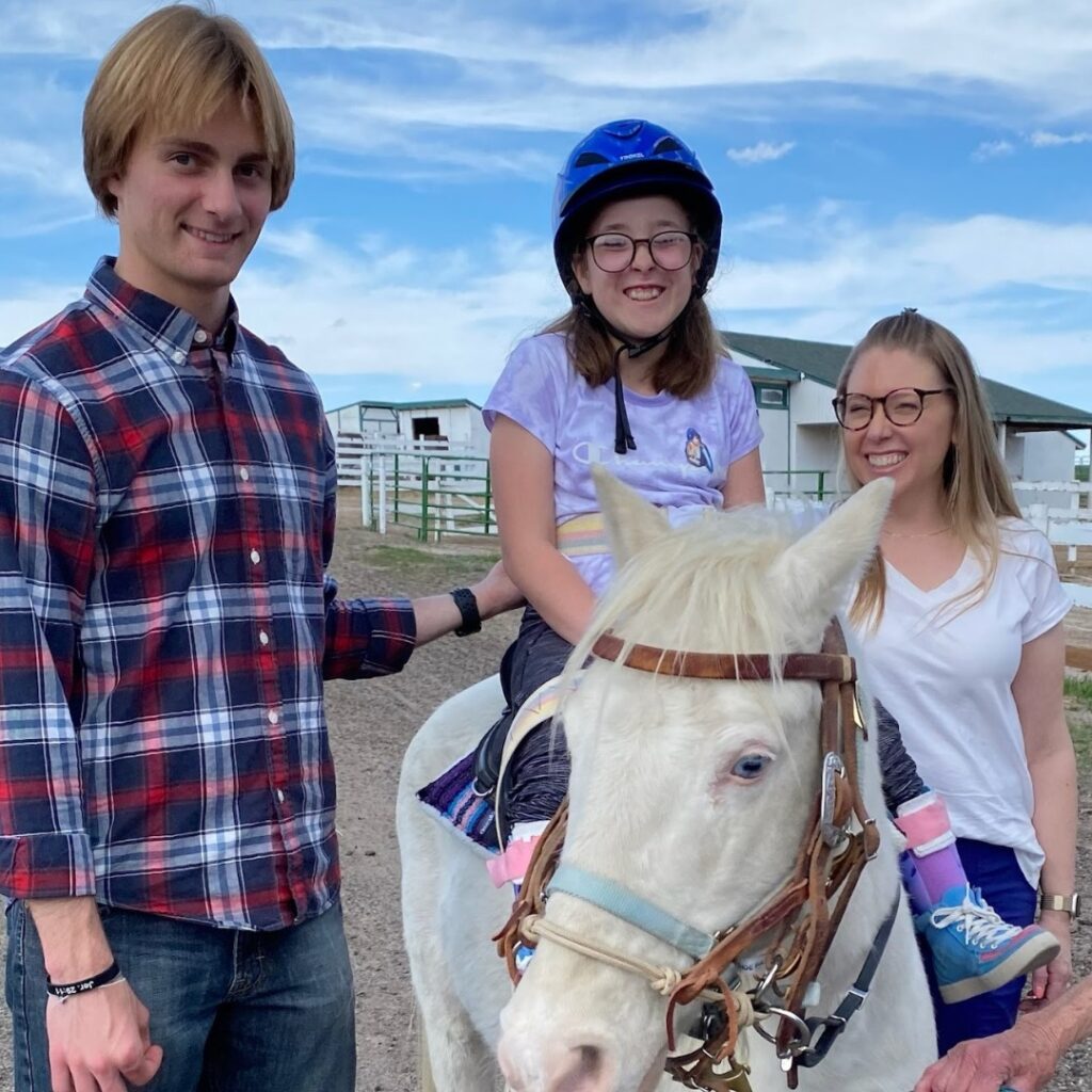 A young girl wearing a blue helmet sits on a white horse, smiling, with a man in a plaid shirt and a woman in glasses and a white shirt beside her at a ranch on a sunny day—this is Chloes Story.