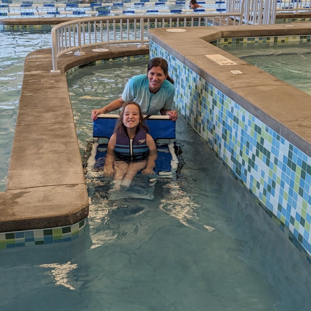 A smiling girl in a life vest sits on a blue flotation device in a pool, supported by an adult from behind. They are both in a narrow, tiled section of an indoor swimming pool, showing a joyful moment from Chloes Story.