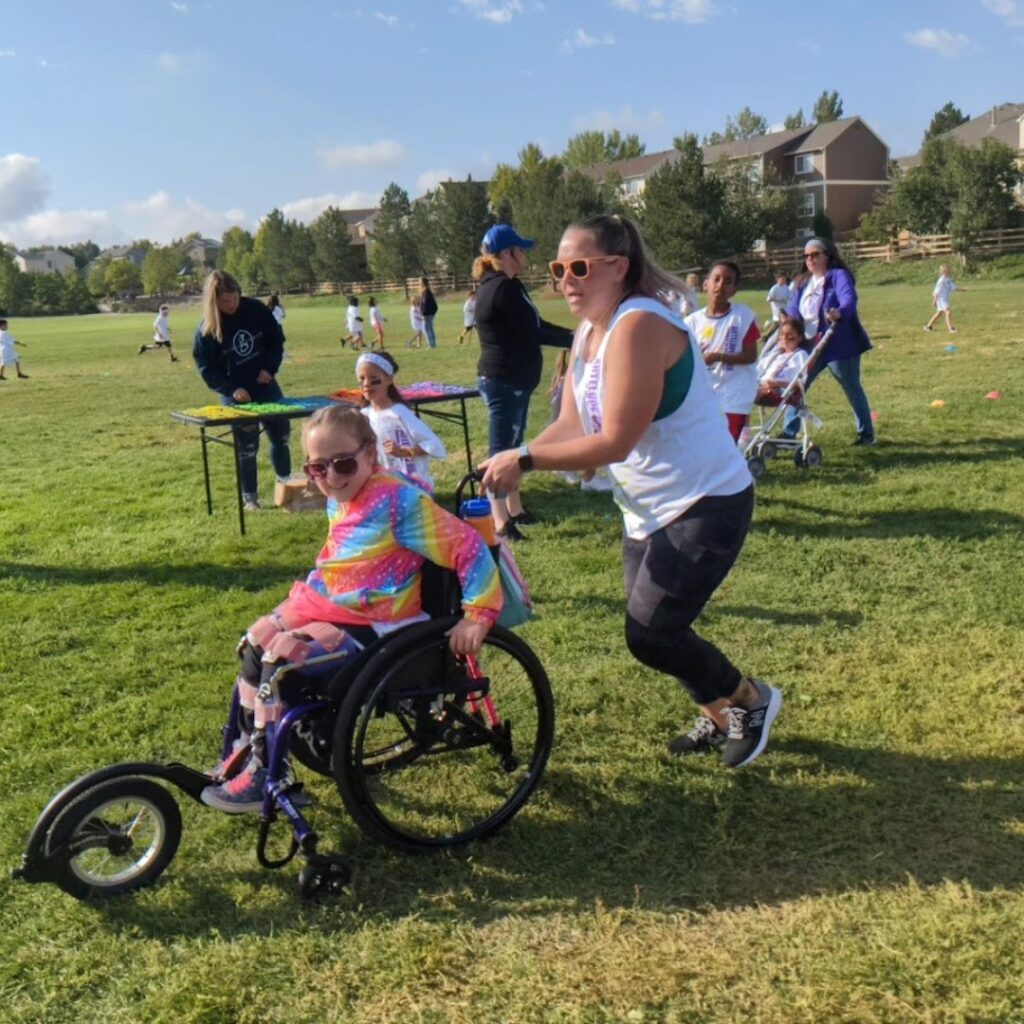 A woman pushes a smiling young girl in a wheelchair across a grassy field during an outdoor event, sharing Chloes Story. Other children and adults are nearby, enjoying the sunny day and participating in activities.