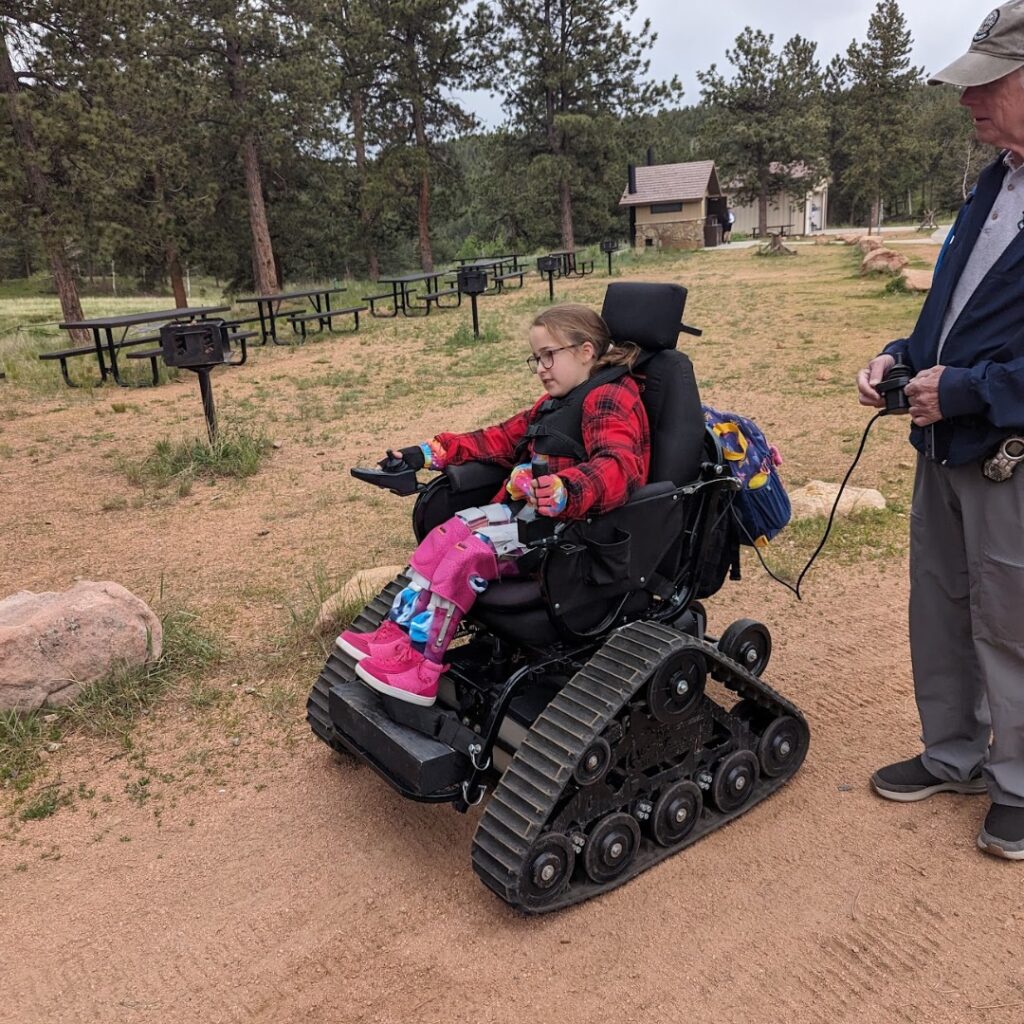 A young girl in a red plaid jacket sits in an all-terrain wheelchair with tank-like tracks on a dirt path surrounded by trees, while an adult stands nearby holding a remote—capturing a moment from Chloes Story.