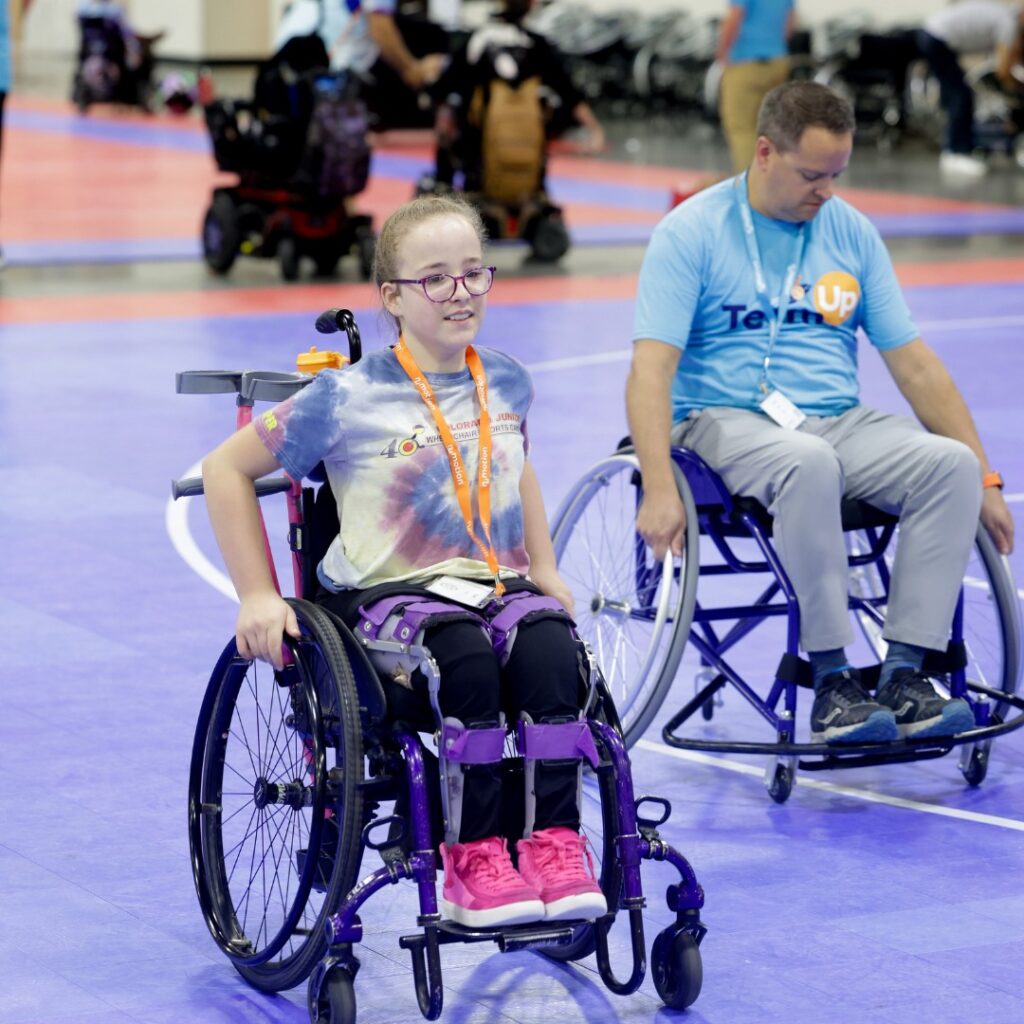 Chloes Story unfolds as a young girl in a tie-dye shirt and pink shoes and a man in a blue shirt participate in an indoor sports event on a purple court, with other participants visible in the background.