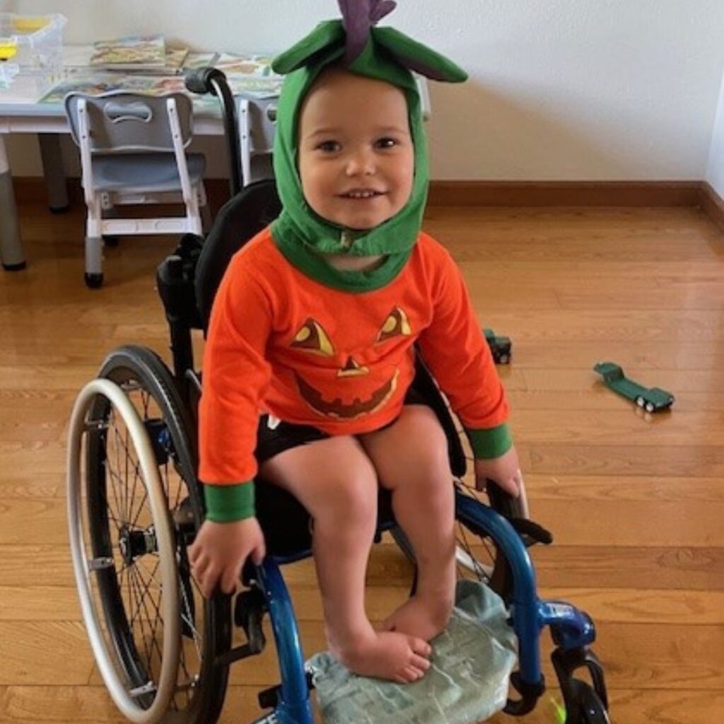 A young child in a wheelchair smiles, wearing a pumpkin-themed costume with a green hood and orange shirt featuring a jack-o’-lantern face. Indoors on a wooden floor, this sweet moment is part of Torreys Story.