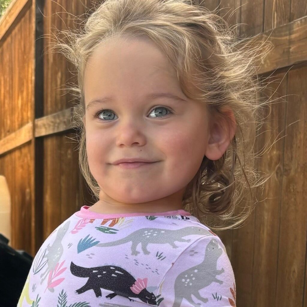 A young child with wavy blonde hair and blue eyes smiles while standing outside near a wooden fence. The child, featured in Torreys Story, is wearing a shirt decorated with colorful animals and plants.