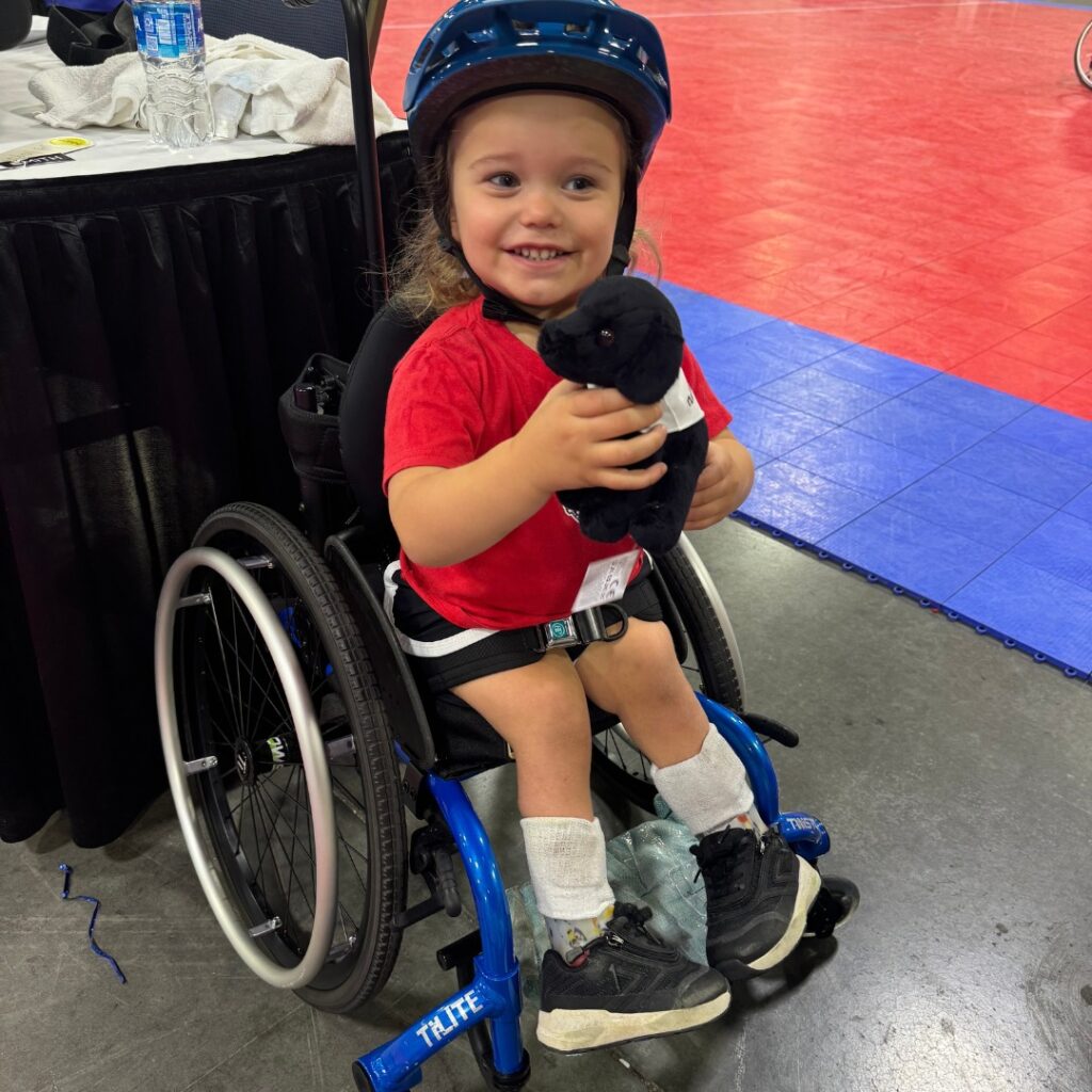 A young child wearing a blue helmet and red shirt smiles while sitting in a wheelchair, holding a small black stuffed animal. The scene, part of Torreys Story, takes place at an indoor sports venue with blue and red mats.