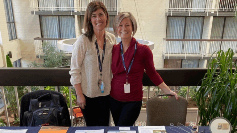 Two women stand and smile behind a display table with pamphlets and materials in a brightly lit indoor space filled with plants and balconies, sharing Alexiss Story.