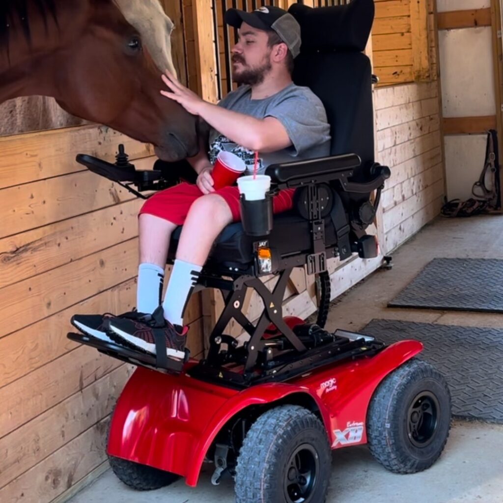 Quintens Story: A man in a red electric wheelchair reaches out to gently touch a brown horse’s face in a wooden stable, smiling as the horse leans forward toward him.
