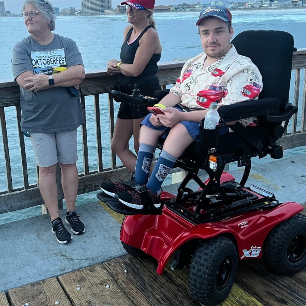 Three people on a pier by the water: one man in a red electric wheelchair, wearing a Cars-themed shirt and cap, and two women stand nearby. All look toward the water, with tall buildings in the background—part of Quintens Story.