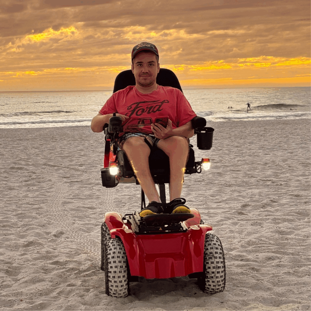 Quintens Story: A man in a red motorized wheelchair sits on a sandy beach at sunset, holding a phone. The orange, cloudy sky hangs over the ocean, with a few people in the background.