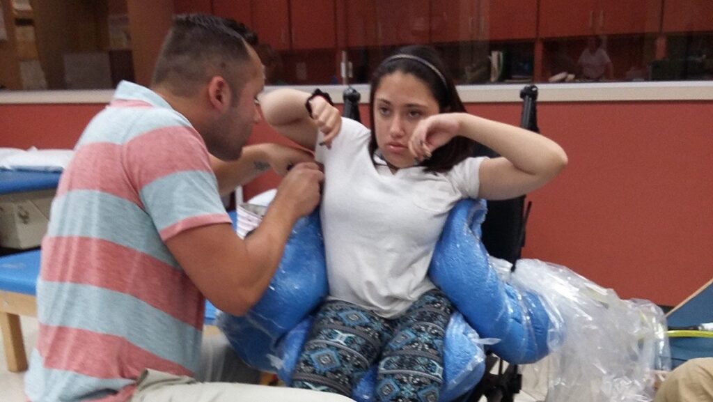 A young woman sits in a wheelchair with blue support cushions, raising her arms, while a man in a striped shirt kneels beside her, assisting her with an exercise in a therapy setting to support mobility and independence.