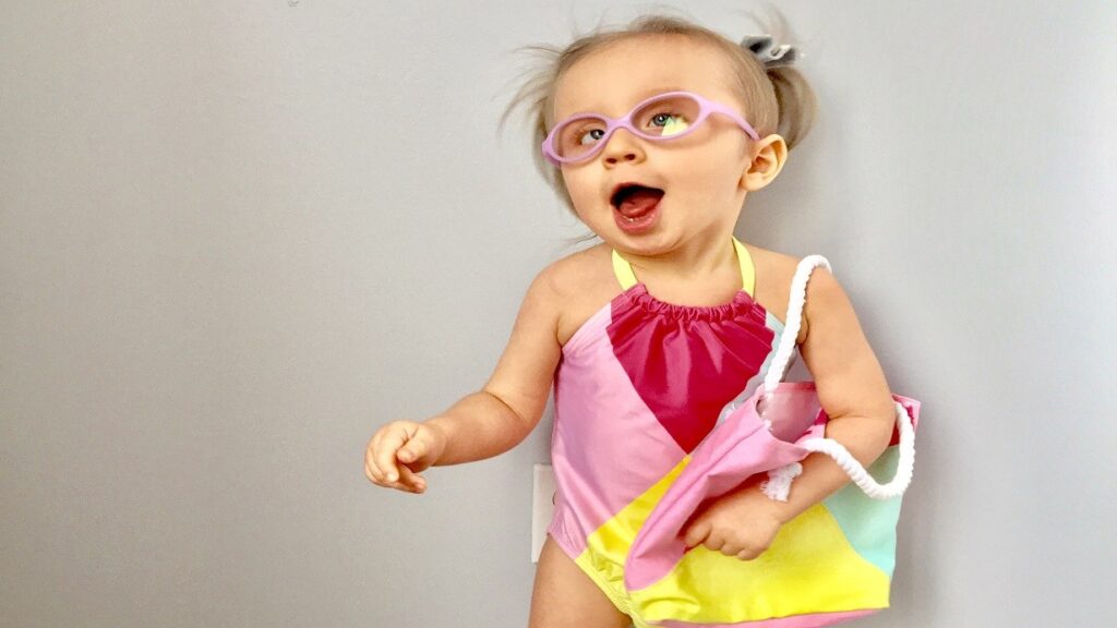 A smiling toddler in pink glasses and a colorful swimsuit holds a matching bag. She stands against a plain light-colored wall, her mouth open in delight, with her hair styled in playful pigtails.