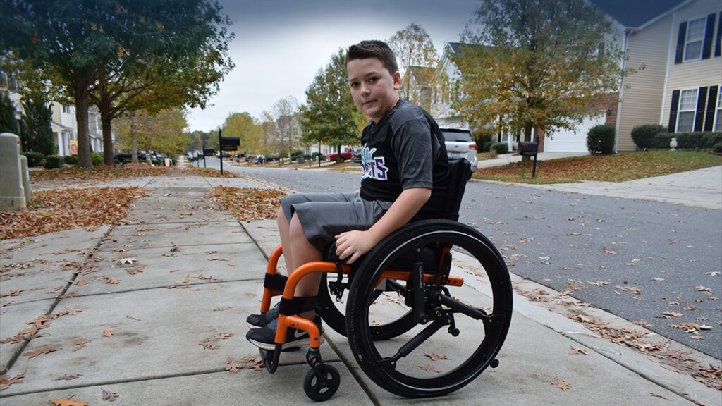 A young boy using a wheelchair sits on a suburban sidewalk lined with autumn leaves and houses, looking at the camera. The quiet street features parked cars and trees with vibrant orange and yellow foliage.