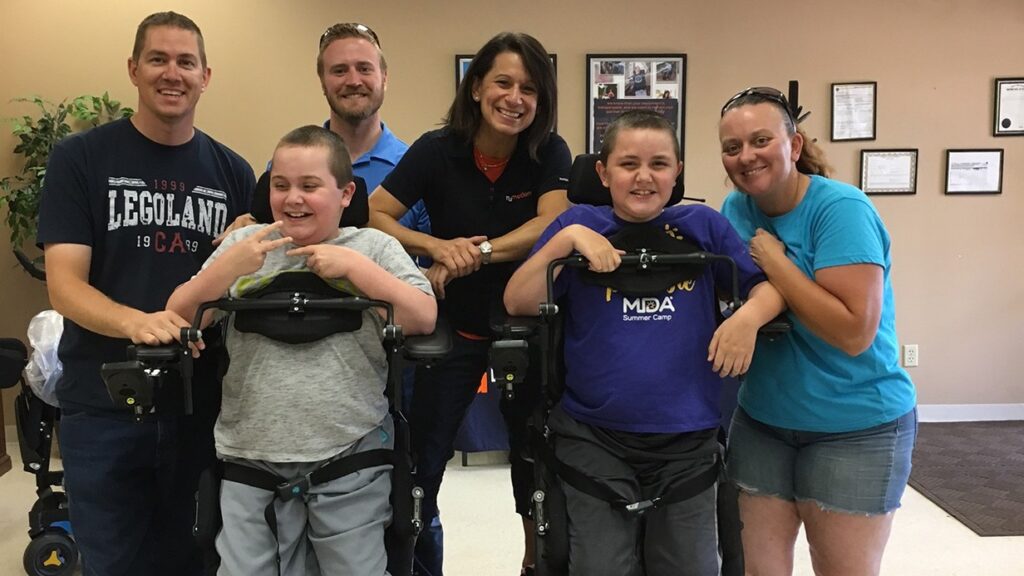 A group of six people, including two children using mobility devices, smile and pose together indoors. Four adults stand behind them, all looking cheerful and supportive.