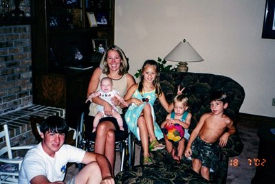 A woman sits on a chair holding a baby, surrounded by four smiling children in a cozy living room. Two kids sit on an armchair, one with a ball, while another boy sits shirtless on the floor.