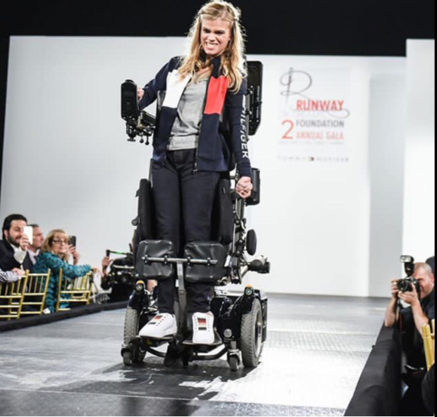 A smiling woman uses a robotic exoskeleton wheelchair to walk down a runway during a fashion show, as photographers and audience members watch and take photos of the innovative mobility technology in action.