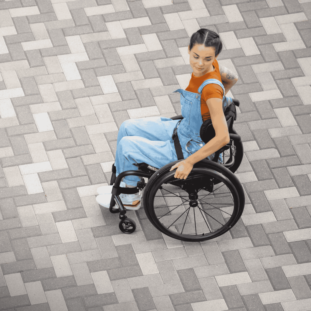 Woman in overalls seated in an ultra-lightweight manual wheelchair on a backdrop of herringbone brick flooring.