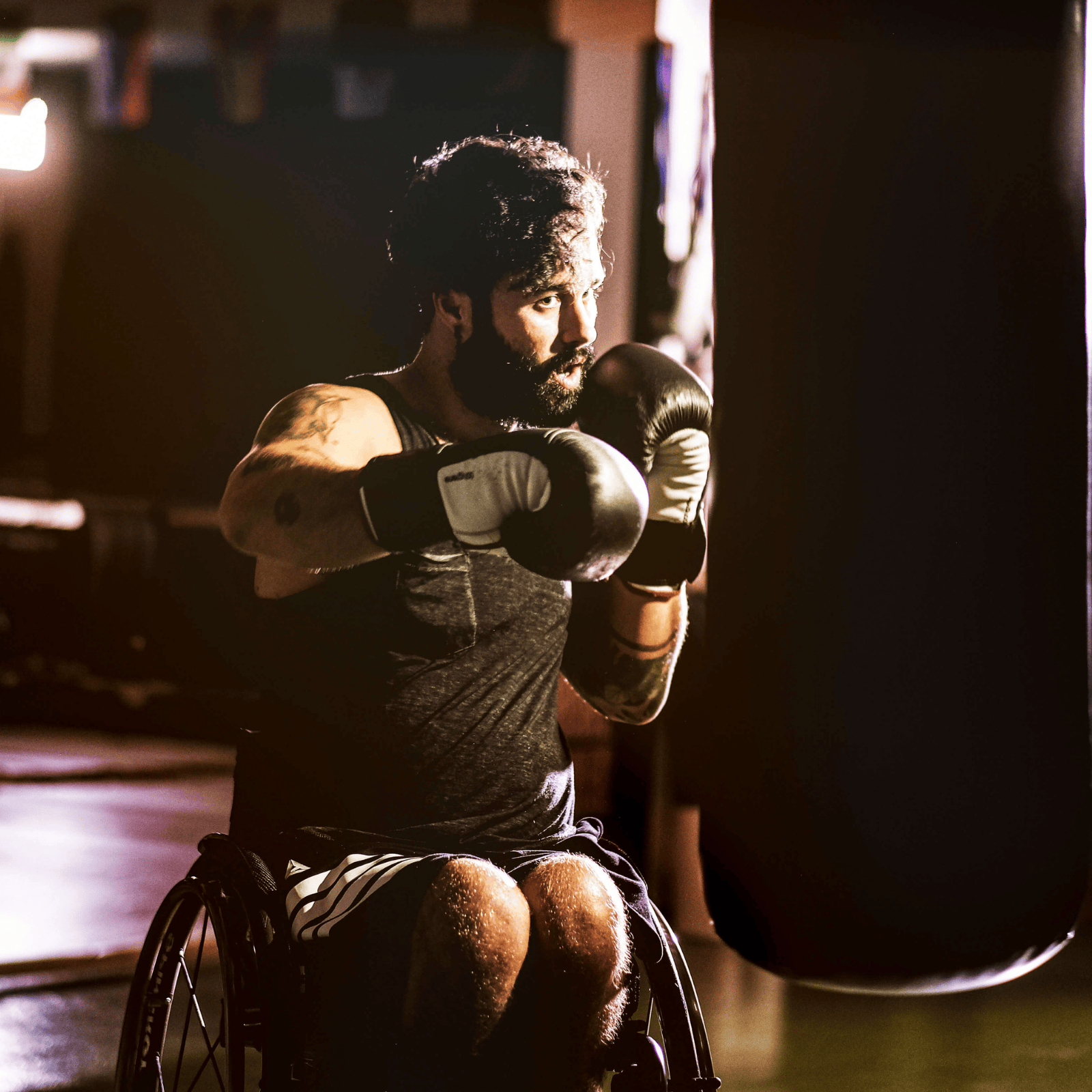 A focused man in a wheelchair trains with boxing gloves, punching a heavy bag in a dimly lit gym. He has a beard, tattoos, and wears athletic gear.