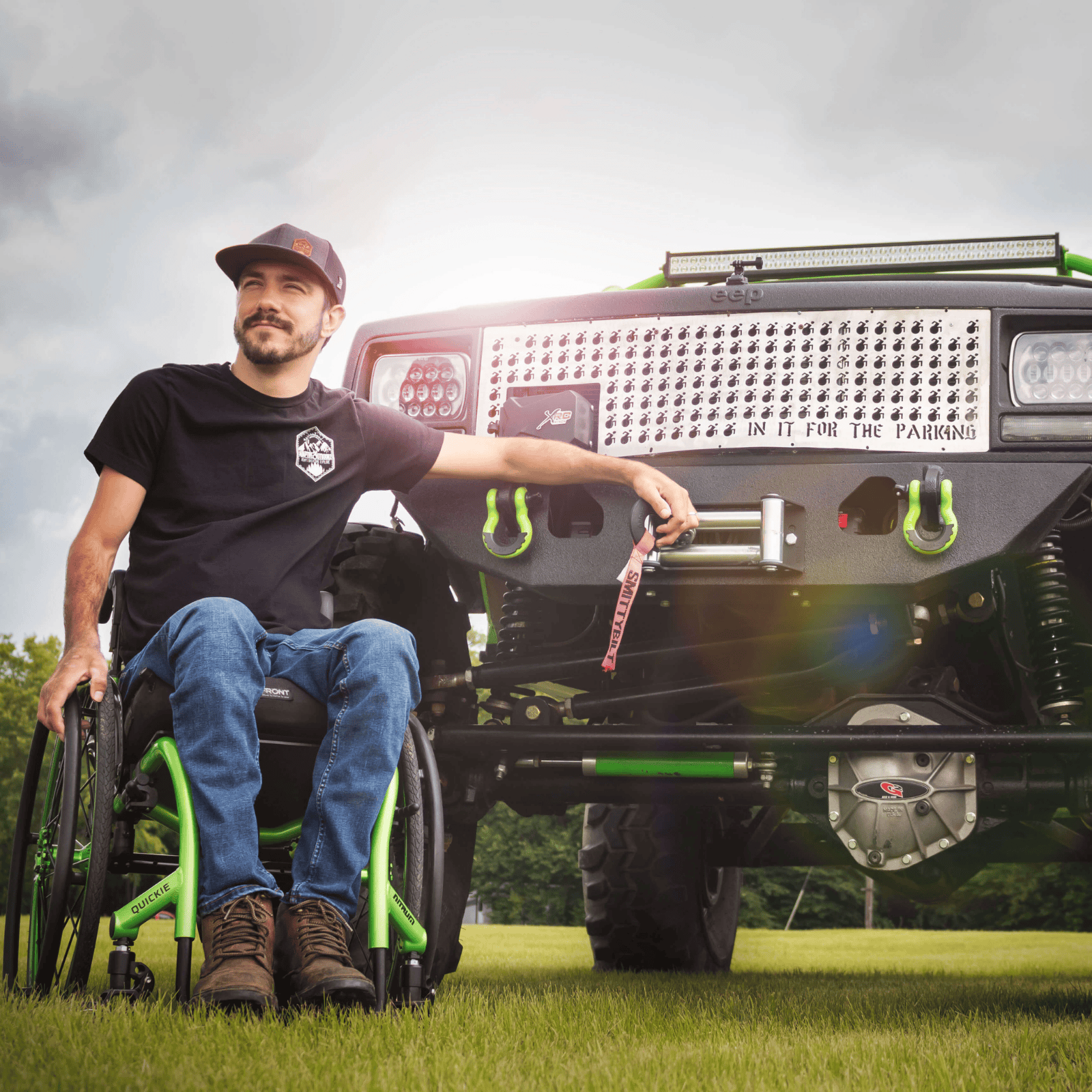 A man in a wheelchair poses confidently next to a modified off-road Jeep. The Jeep’s grill features a pattern of wheelchair symbols and the phrase “In It for the Parking.”