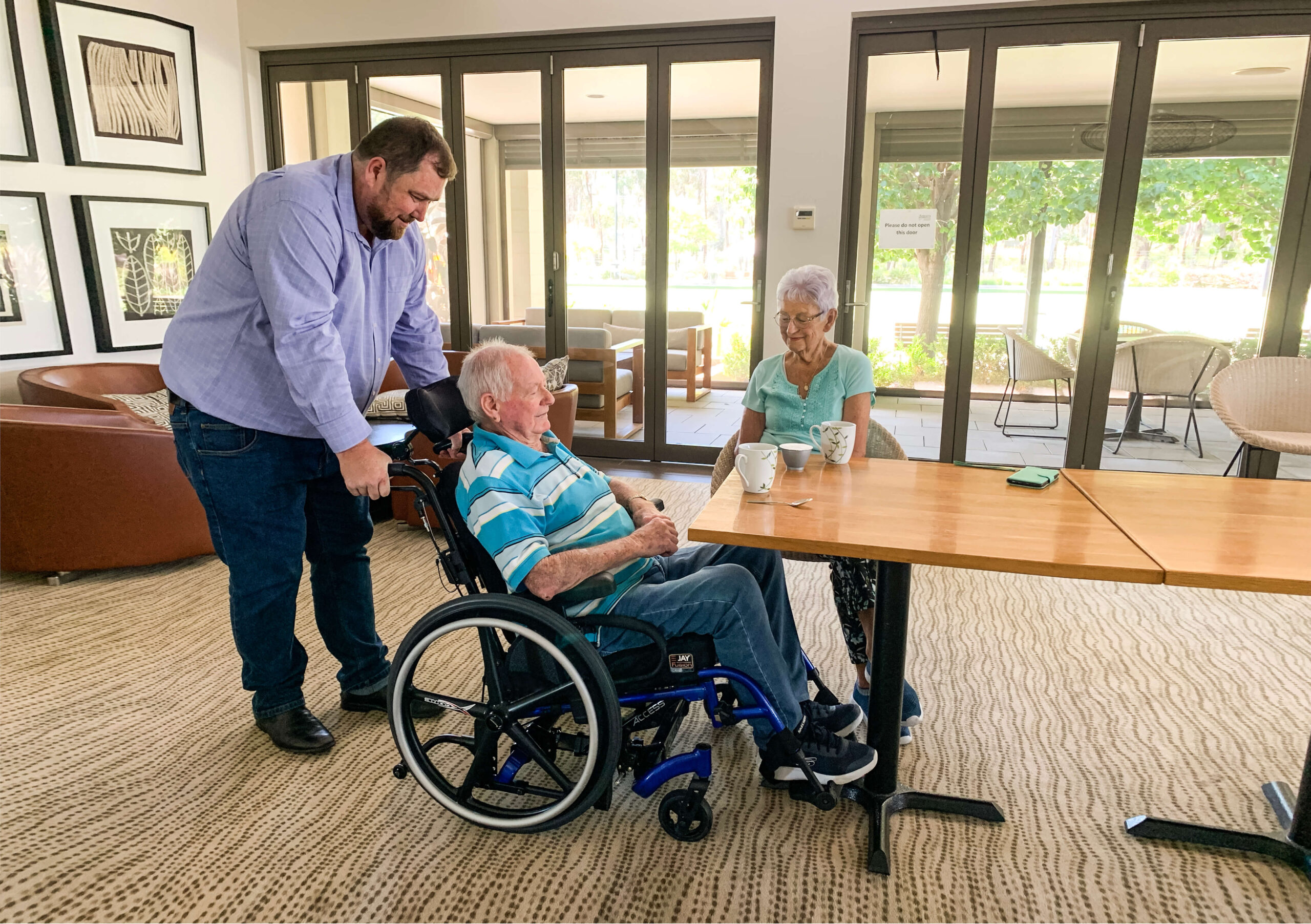 A man stands beside an elderly man in a Power Tilt Wheelchair and an elderly woman seated at a table, all smiling and chatting in a bright, modern room with large windows.