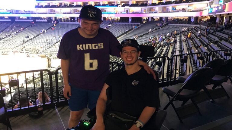 Two men wearing Sacramento Kings shirts and hats pose and smile inside a basketball arena. One, standing with his arm around the other, shares a proud moment. The seats and court set the scene for Adams Story.