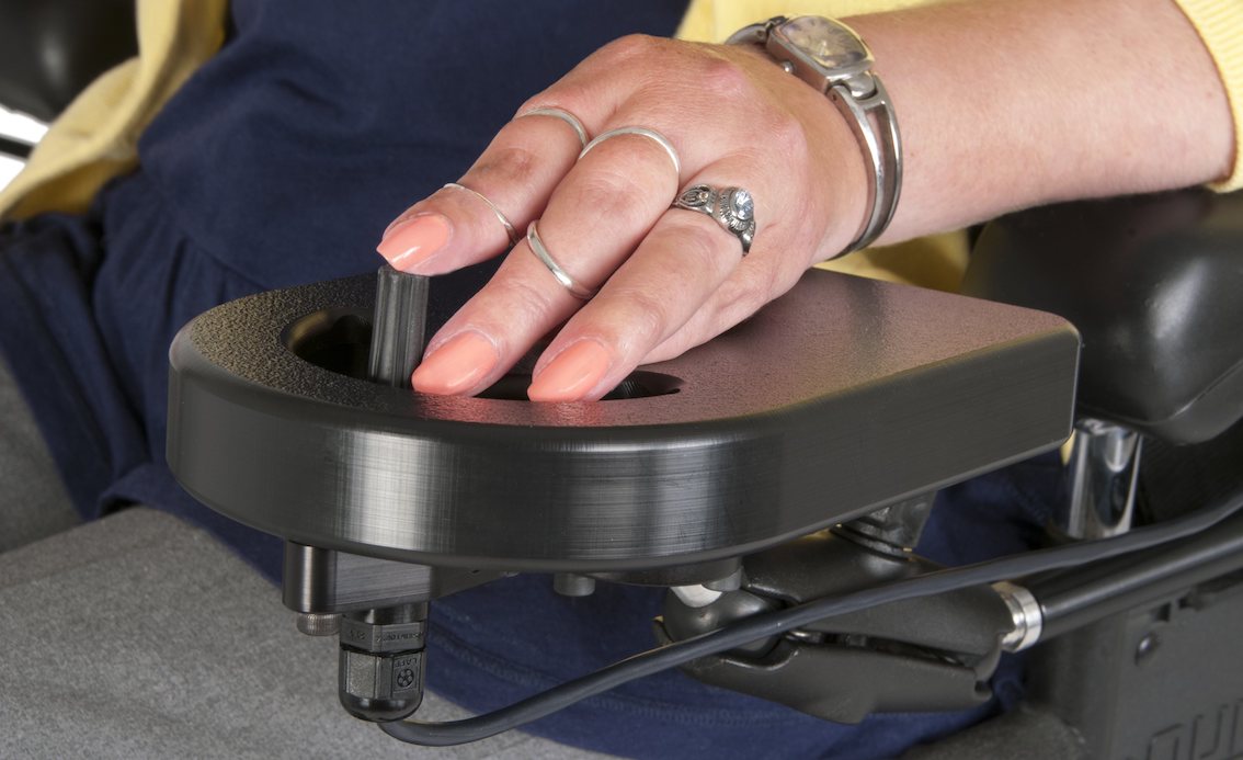 A person wearing rings and a watch uses their hand to operate a joystick control on a wheelchair, demonstrating alternative access methods for improved independence.