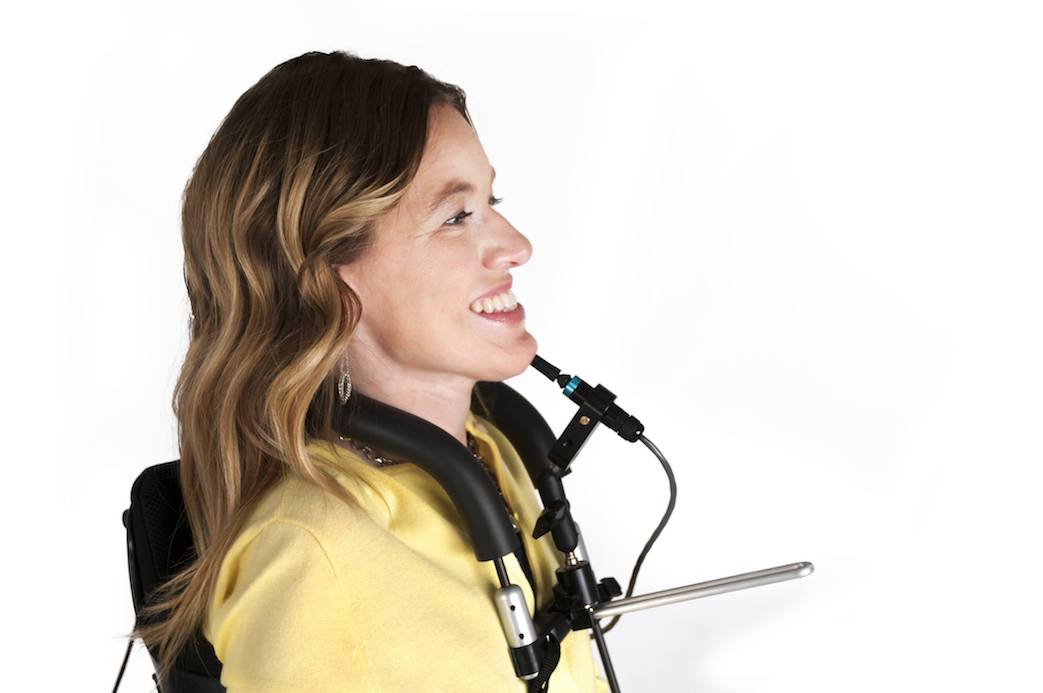 A woman with light brown wavy hair, wearing a yellow top, smiles while using a wheelchair equipped with a headrest, a mouth-operated joystick, and Alternative Access Controls. The background is plain white.