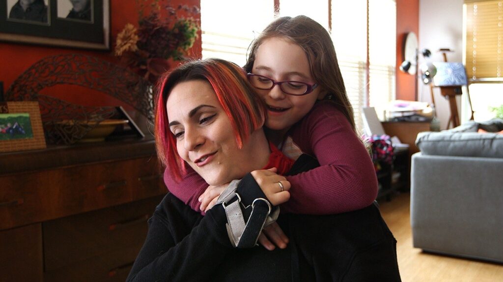 A young girl with glasses and long hair hugs a smiling woman with short, red-streaked hair from behind in a cozy, sunlit living room. The woman has a brace on her right arm, capturing a warm moment from Allainas Story.