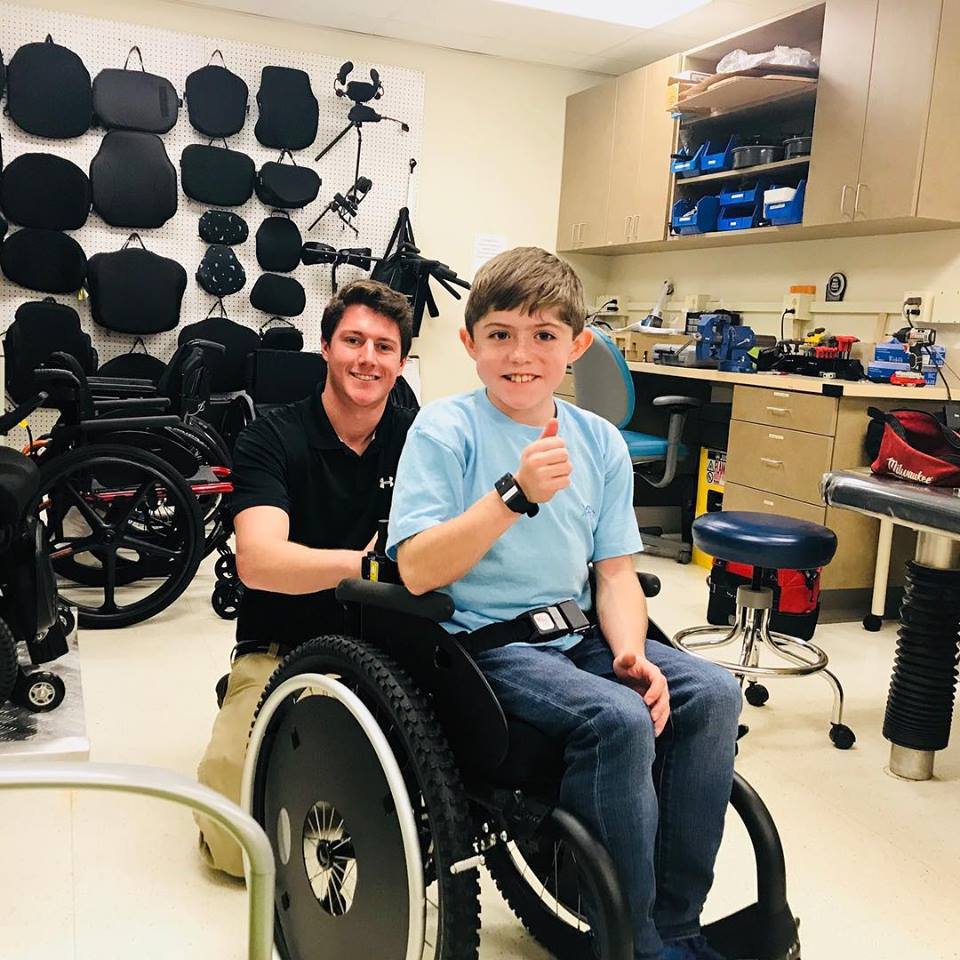 A smiling boy in a wheelchair gives a thumbs up while a man kneels behind him in a workshop filled with mobility equipment and tools. They both look happy in the well-lit workspace.