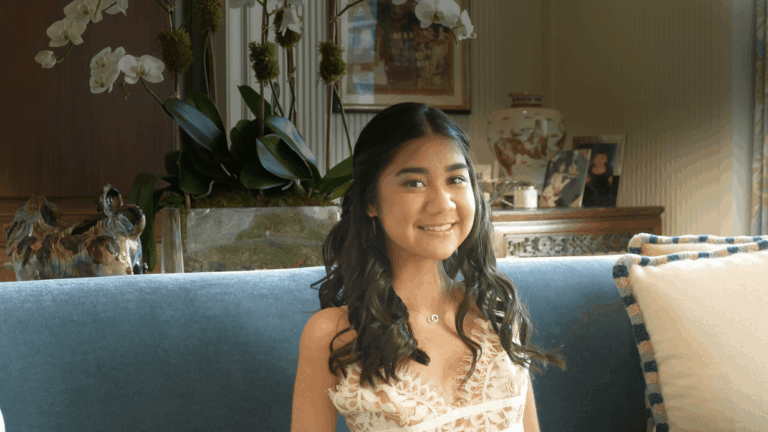 A young woman with long dark hair in loose curls sits on a blue couch, smiling. She wears a white lace dress. Behind her, potted orchids and framed photos add charm to the setting, reflecting Aryas Story.