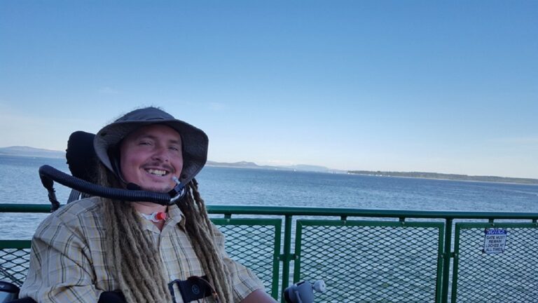 A smiling man with long dreadlocks and a sun hat sits in a power wheelchair on a ferry, with sparkling water and a distant shoreline beneath a clear blue sky.