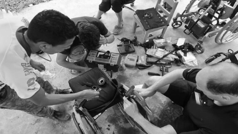 Three men repair a wheelchair outdoors, tools and equipment spread out on the ground; another person stands nearby, partially visible. This scene is part of Mikes Story.